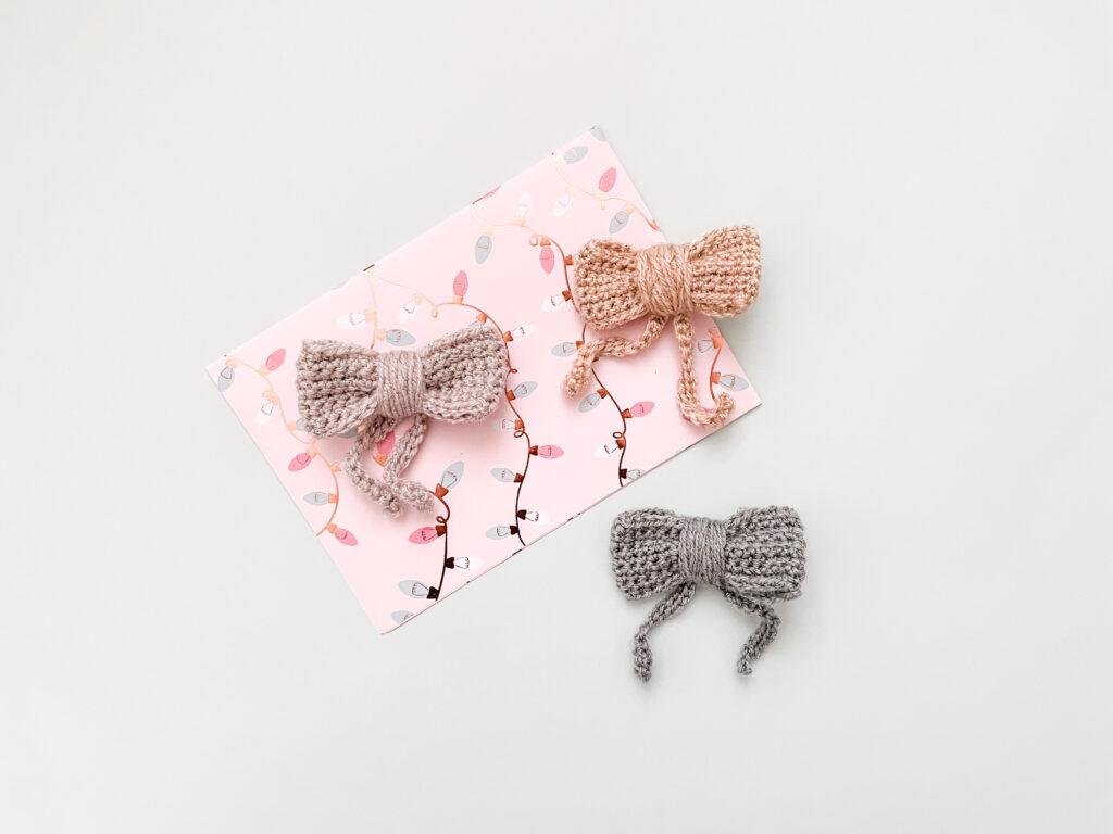 a flat lay image of three crochet bows made in gold, silver and pale green sparkly yarn lay on a pale blue backdrop and pink wrapping paper
