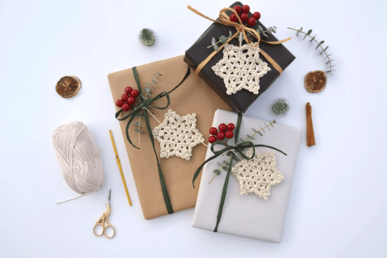A trio of crochet snowflakes in oatmeal coloured yarn adorn Christmas parcels wrapped in neutral wrapping paper. They are decorated with green twine and holly berries. 