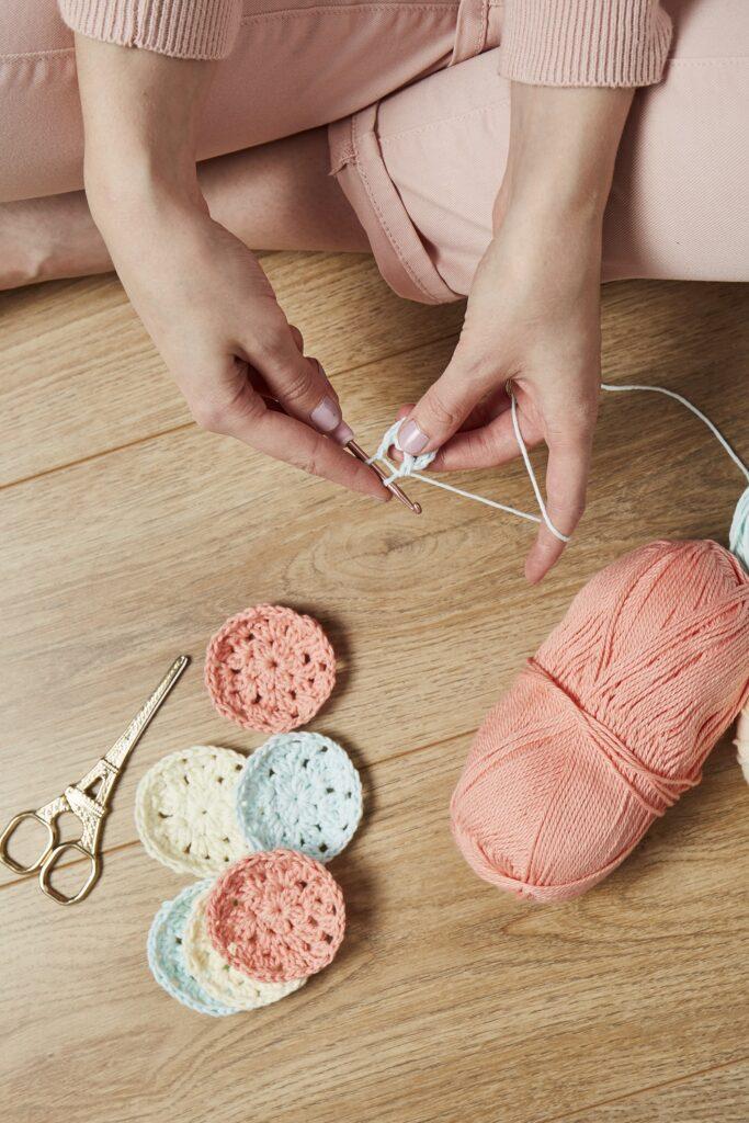 a woman sits on a wooden floor crocheting make-up scrubbies in shades of pink, cream and pale blue. 