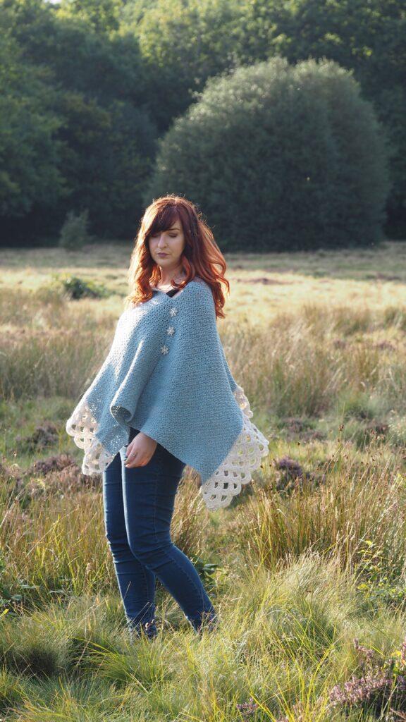 A woman with red hair is standing in moorland. She is wearing a crochet poncho in shades of blue and cream with a lace border. 