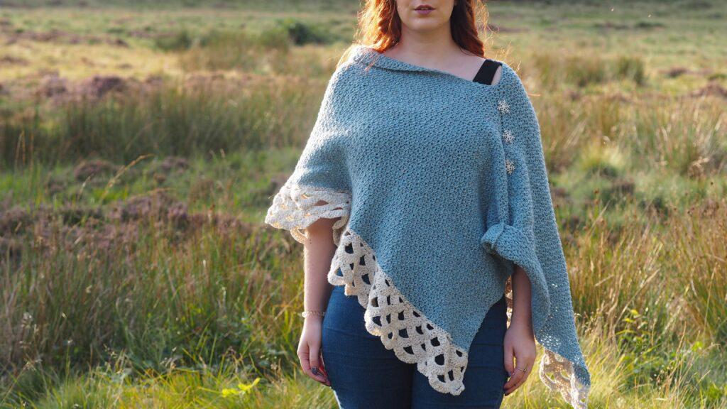 A redheaded woman is wearing a blue and cream crochet poncho with three crystal snowflake buttons. She is standing on moorland.