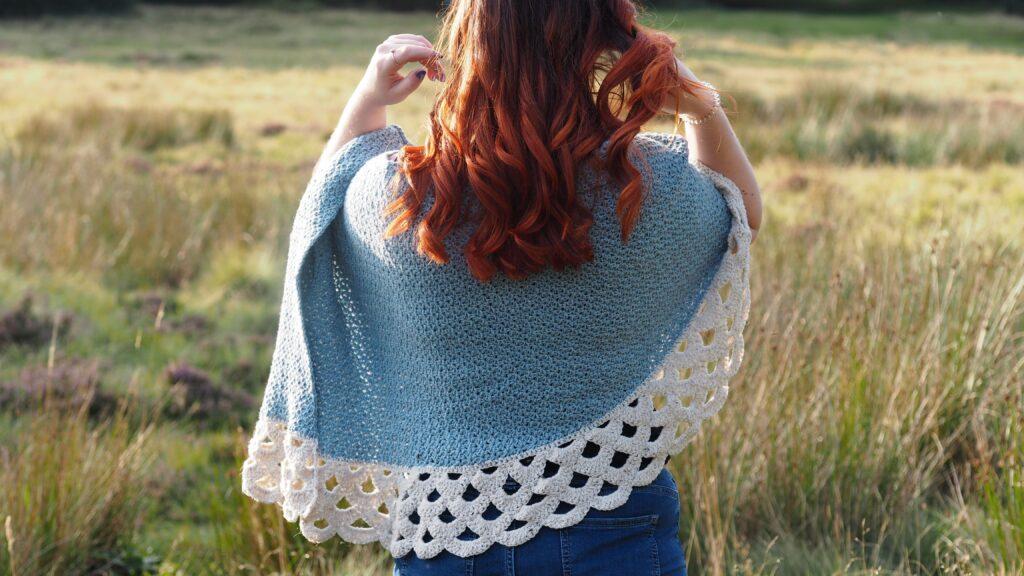 A woman with red hair is standing in moorland. She is wearing a crochet poncho in shades of blue and cream with a lace border. 