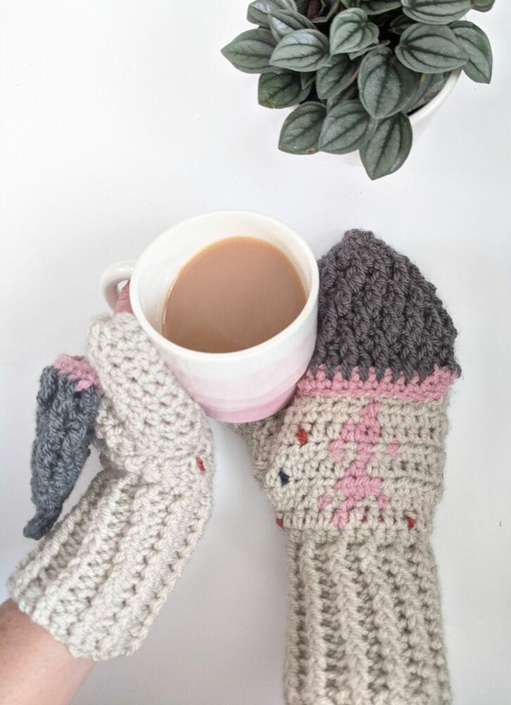 A pair of hands wearing crochet mittens hold a cup of tea. The mittens are alpine-themed in shades of tan, pink and grey.