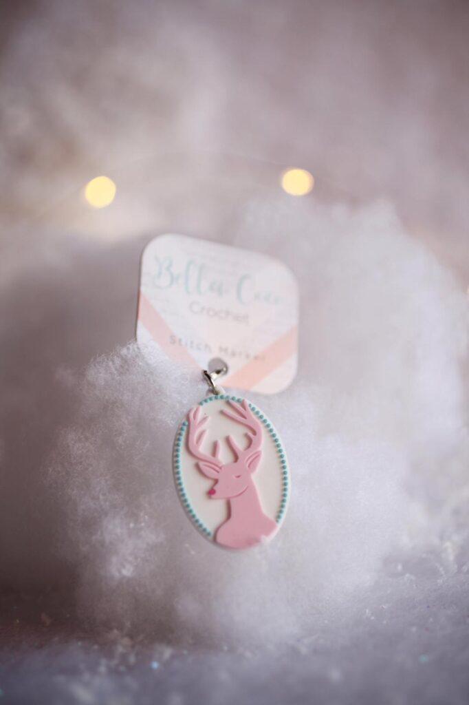 A stitch marker in the shape of a stag's head is displayed. The stag is pink. 