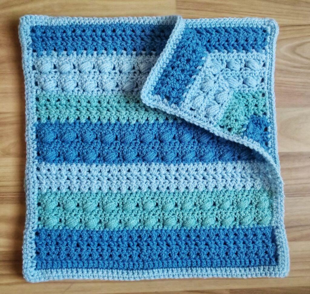 A blue crochet cushion in progress sits on a wooden background. The cushion has different colour and texture stripes in varying shades of blue. 