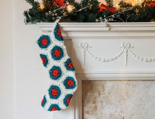 a crochet hexagon stocking made in red, green and cream yarn hanging from a cream mantle with a festive garland draped over the mantle piece.