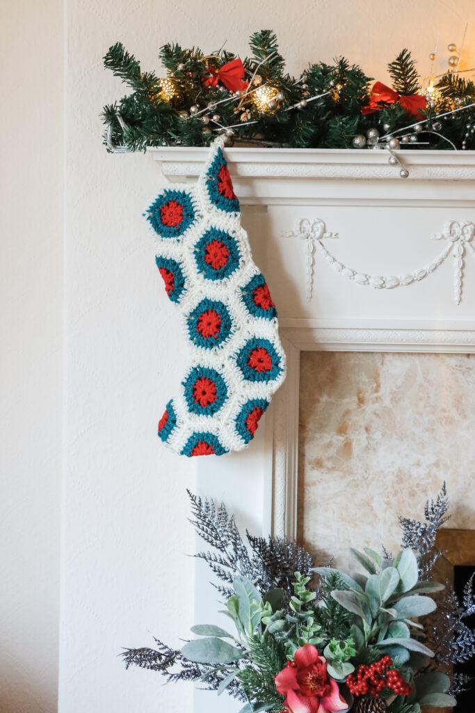 A crochet christmas stocking made of white, green and red hexagons is hanging on a festively dressed white fireplace.