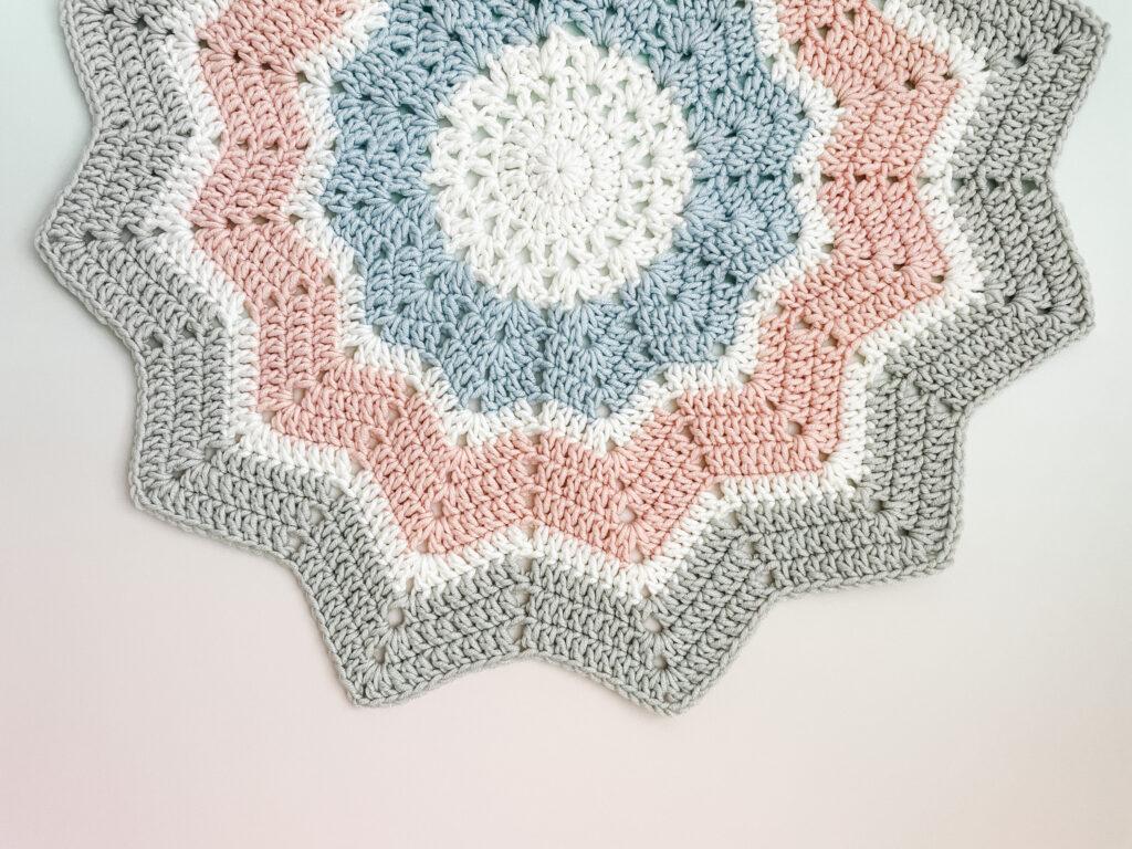 12 Point Star crochet blanket in grey, white, pink and blue yarn as a flat lay on an ombre pink and white background.