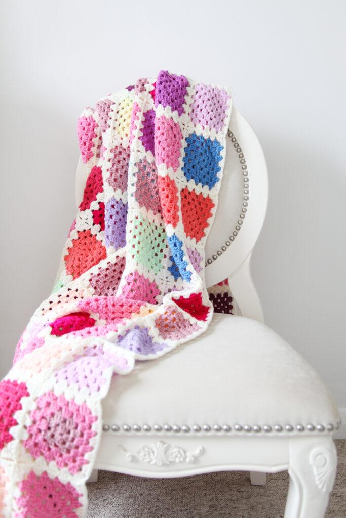 a granny square blanket Mae of multi colours granny squares draped over a white chair against a white wall. 
