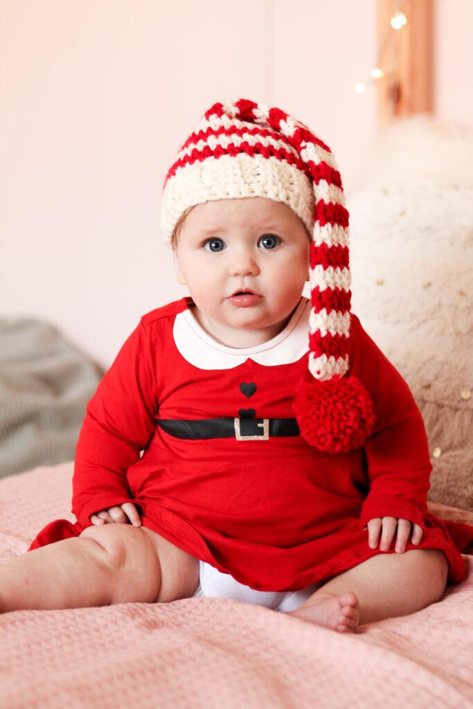 baby sat on a bed with pale pink bedding wearing a red Christmas dress and a crochet baby elf hat.