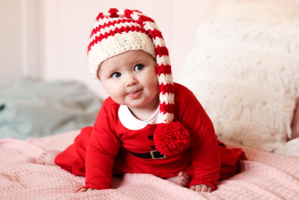 baby sat on a bed with pale pink bedding wearing a red Christmas dress and a crochet baby elf hat. 