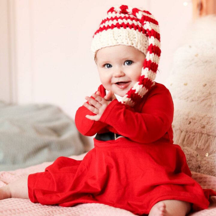 baby sat on a bed with pale pink bedding wearing a red Christmas dress and a crochet baby elf hat.