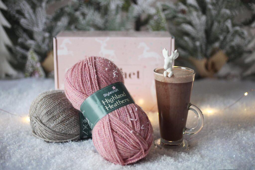 A pair of yarn balls, one in soft pink one in soft brown, sit crossed on a snowy background next to a mug of hot chocolate.