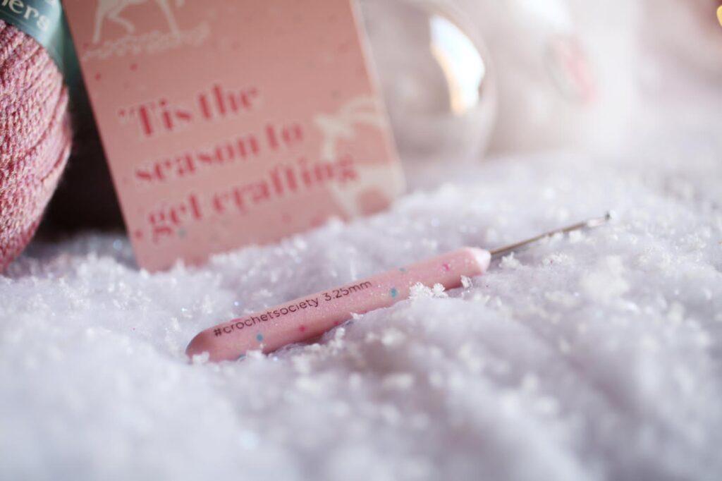 A sparkly pink crochet hook is displayed on a snowy backdrop.