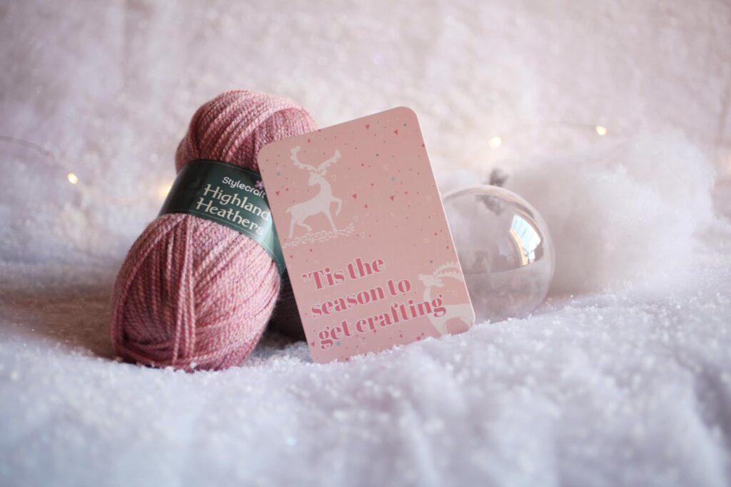 A trio of crochet items are displayed, a ball of pink yarn, a card saying, "Tis the season to get crafting" and a clear bauble. 