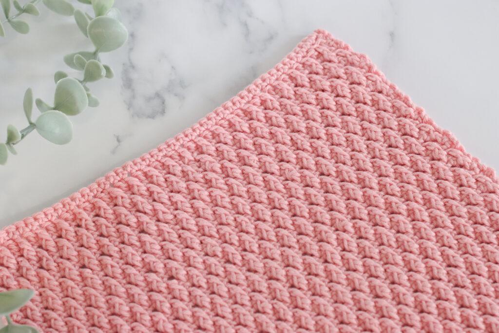 a pink crochet swatch of the alpine stitch on a marbled backdrop and foliage to the left of the image. 