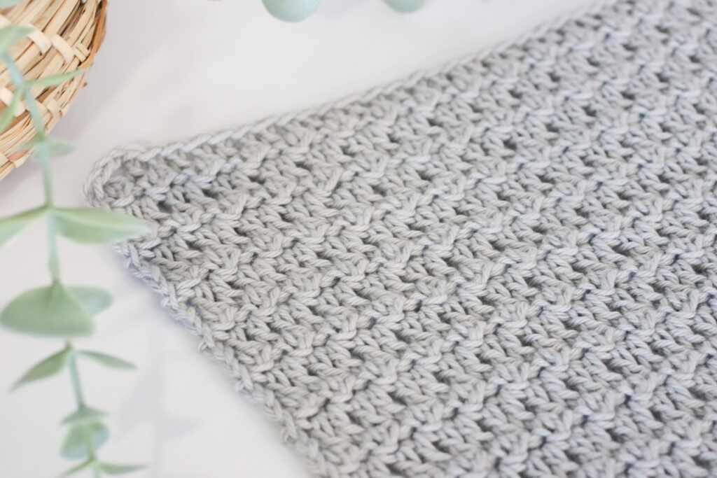Floret crochet stitch swatch in grey yarn on a white background next to a plant in a wooden basket.