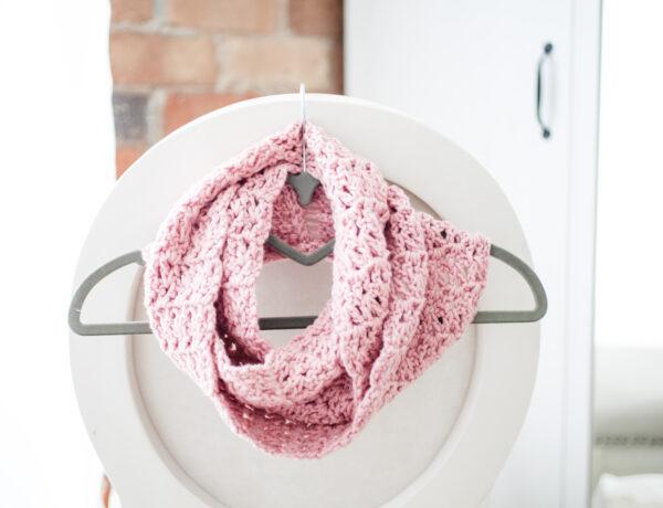 a pink infinity scarf wrapped around a grey coat hanger hooked on the back of a white chair
