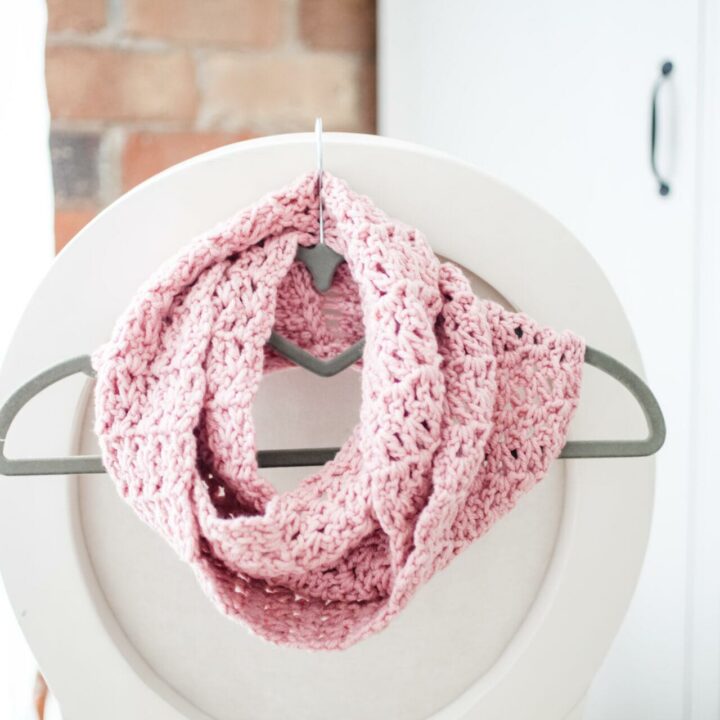 a pink infinity scarf wrapped around a grey coat hanger hooked on the back of a white chair