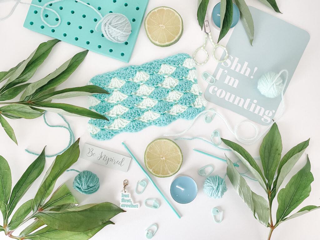 flay lay of blue and green items on a white background. Items include a crochet shell stitch swatch, a crochet hook, yarn, stitch markers, slices of line, foliage, a blue tea light candle and a postcard that says shh I'm counting. 