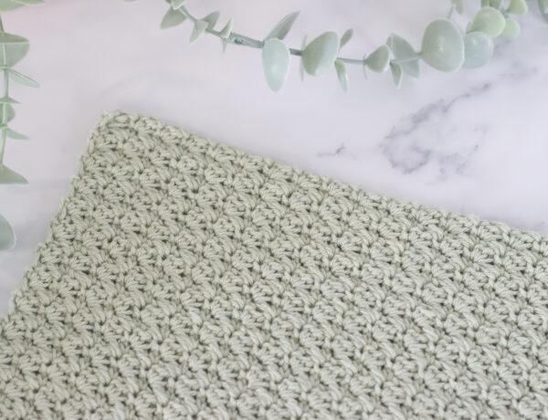 The suzette stitch as a swatch in sage green yarn on a marble background with a plant.