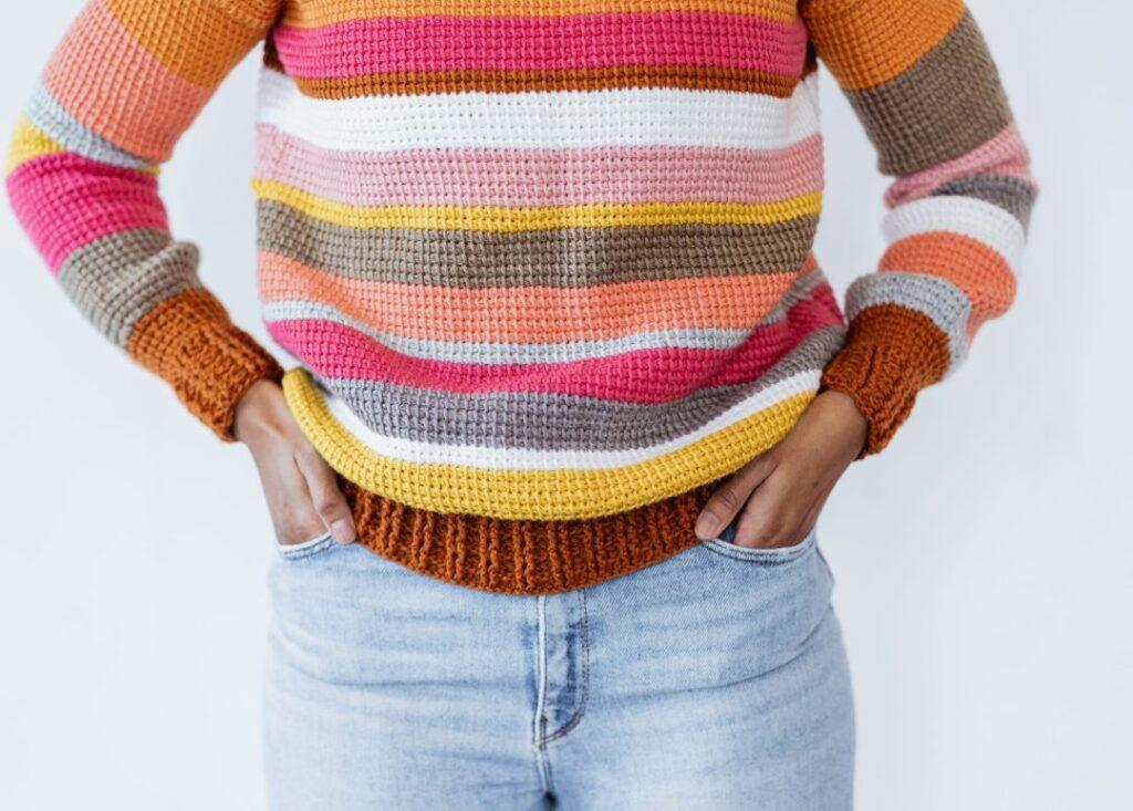A woman wears a pair of blue jeans teamed with a bright-toned crochet sweater. The jumper is made from pink, orange, yellow and neutral stripes. 