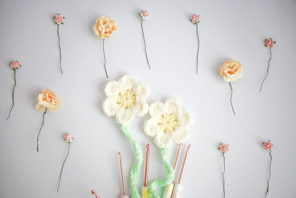 A pair of crocheted daisies sit on a white background alongside paper roses in shades of pale pink and yellow. 