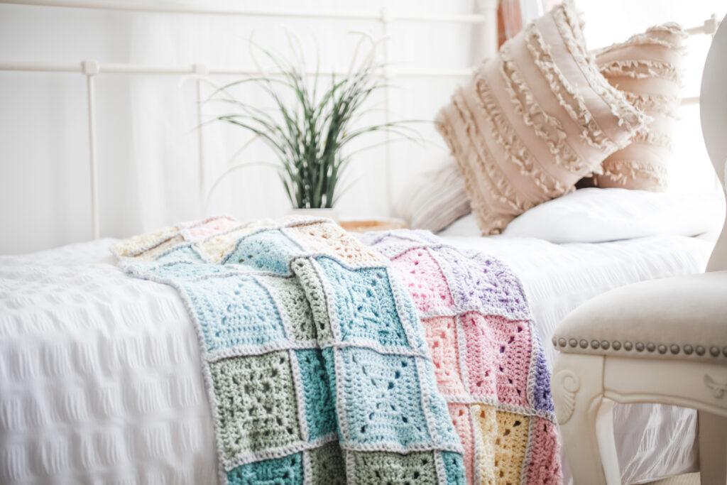 A pastel coloured blanket sits on top of a white bed. The blanket is made from chunky yarn crocheted into decorative squares. 