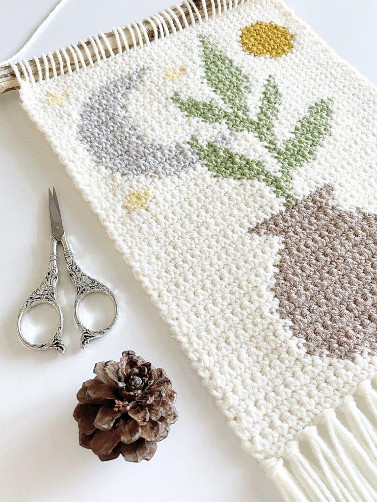 The Oh So Boho wall hanging sits on a white background beside a pair of silver scissors and a pine cone. 