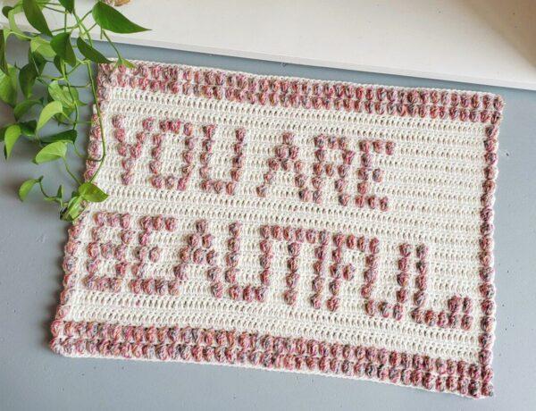 a flat lay image of a textured crochet bath mat on the floor that says You are beautiful within the stitches.
