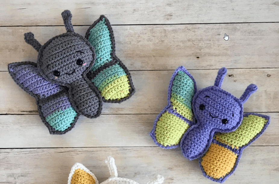 A pair of crochet butterflies sit on a wooden floor. They are made of teal, purple, black and lime yarn. 
