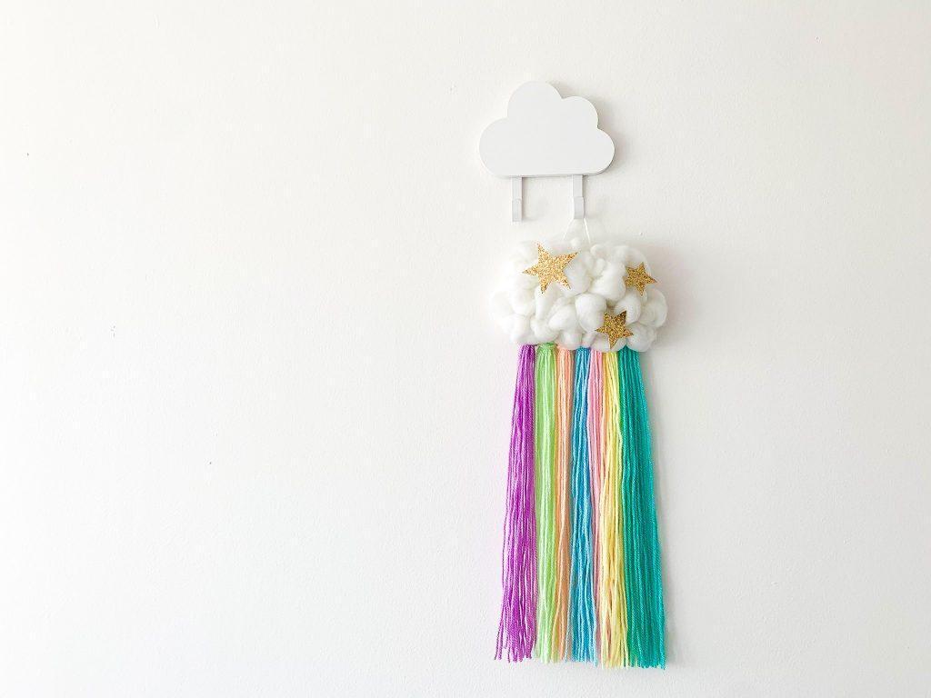 A white cloud made from yarn and studded with gold, glitter stars sits on a white wall. There are streams of pastel rainbow coloured yarn falling from the bottom of the cloud. 