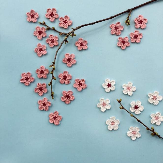 An arrangement of crocheted cherry blossom flowers in various shades of pink are arranged alongside two branches on a blue background. 