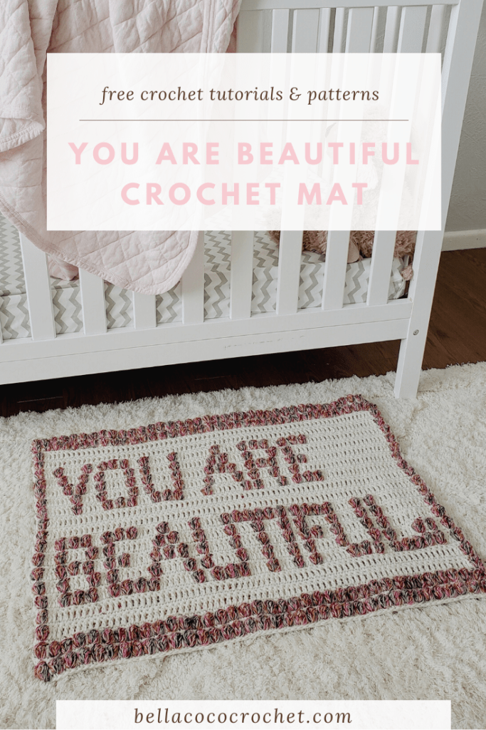 A crochet floor mat sits beside a white crib. The crochet mat has bobble stitches spelling out the words, "You Are Beautiful" in capital letters.