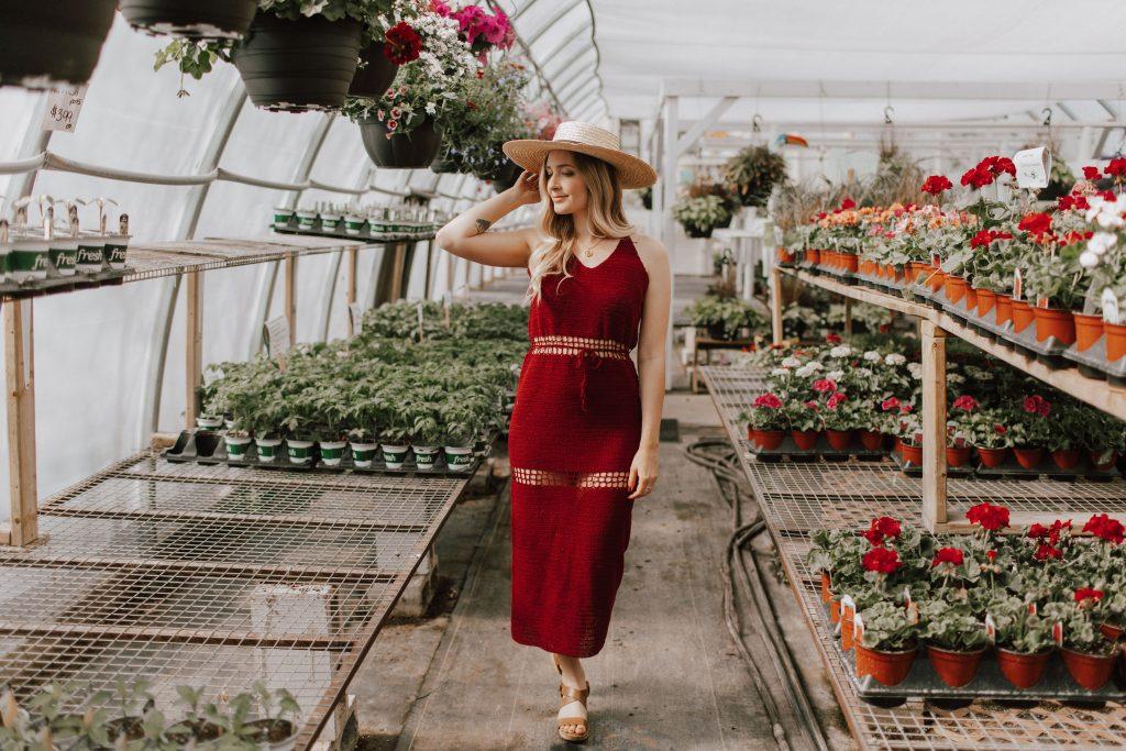 A woman stands in a greenhouse surrounded by flowers. She is wearing a red crochet dress and a straw sunhat. 