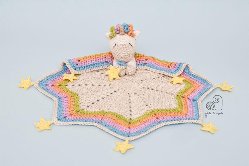 A unicorn theme lovey sits on white background. This crochet toy is made from pastel toned yarns and has stars attached to the blanket base.