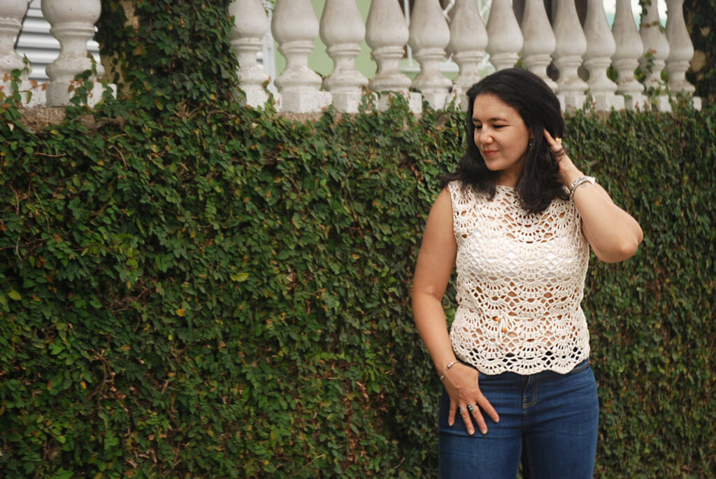 A woman stands to the right of the frame against a deep green hedge. She is wearing a white, lacy crochet top and blue jeans.