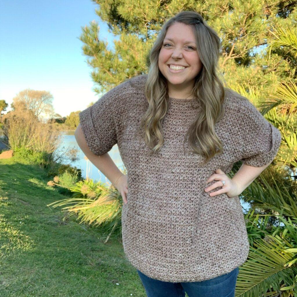 A woman stands surrounded by trees, she is wearing a taupe coloured crochet tee in a slouchy style. She is smiling. 
