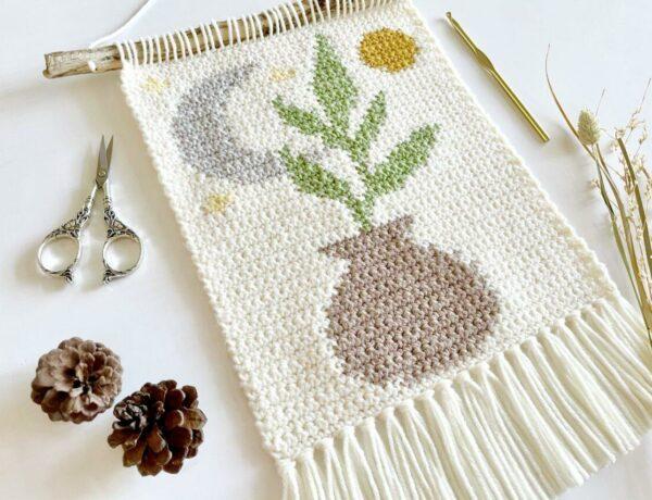 a flat lay image of a crochet wall hanging, the tapestry design features a vase, leaves, a present moon and the sun.