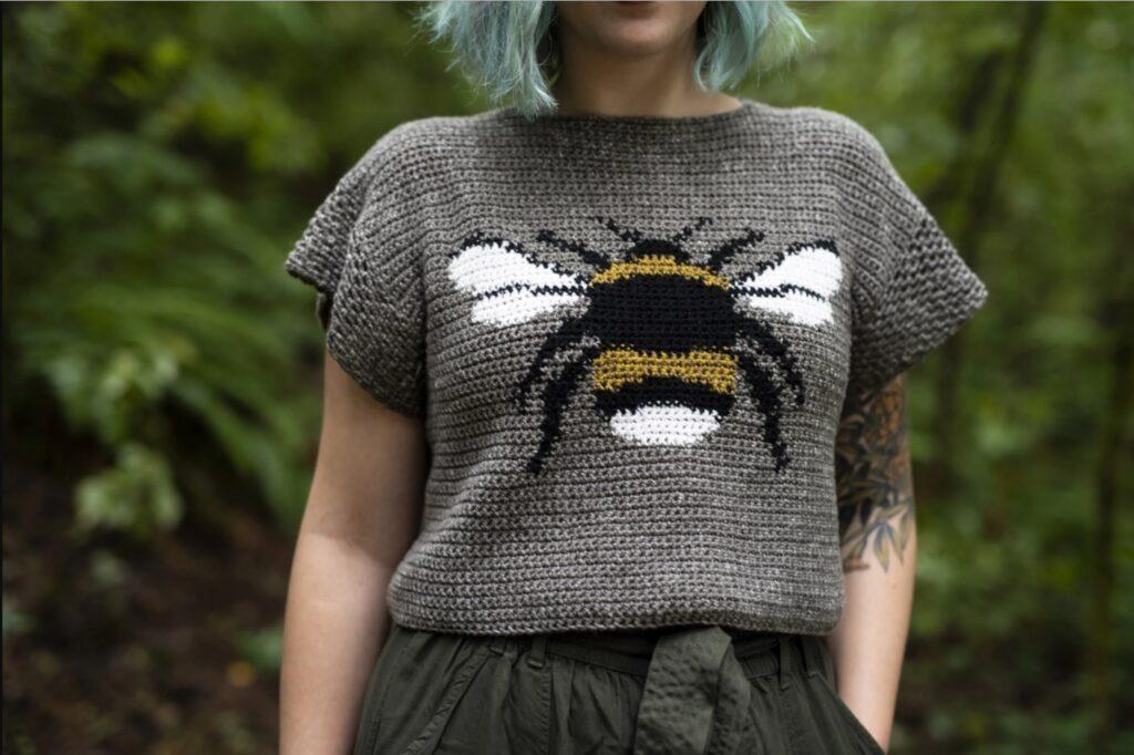 A woman is standing in a forest wearing a brown crochet tee with a large bee motif on the front. 