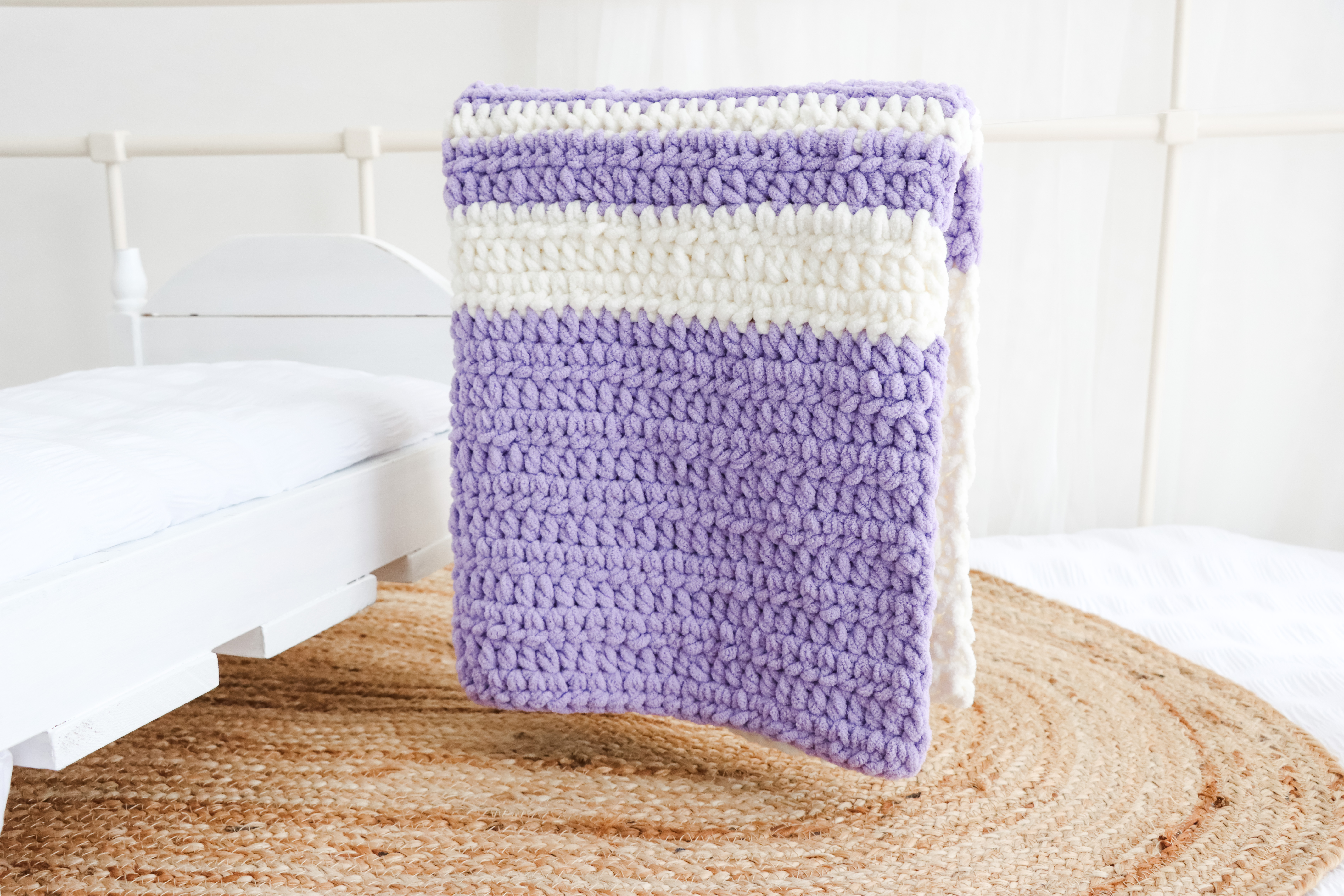 A lilac and white striped blanket is draped over a white bed post.
