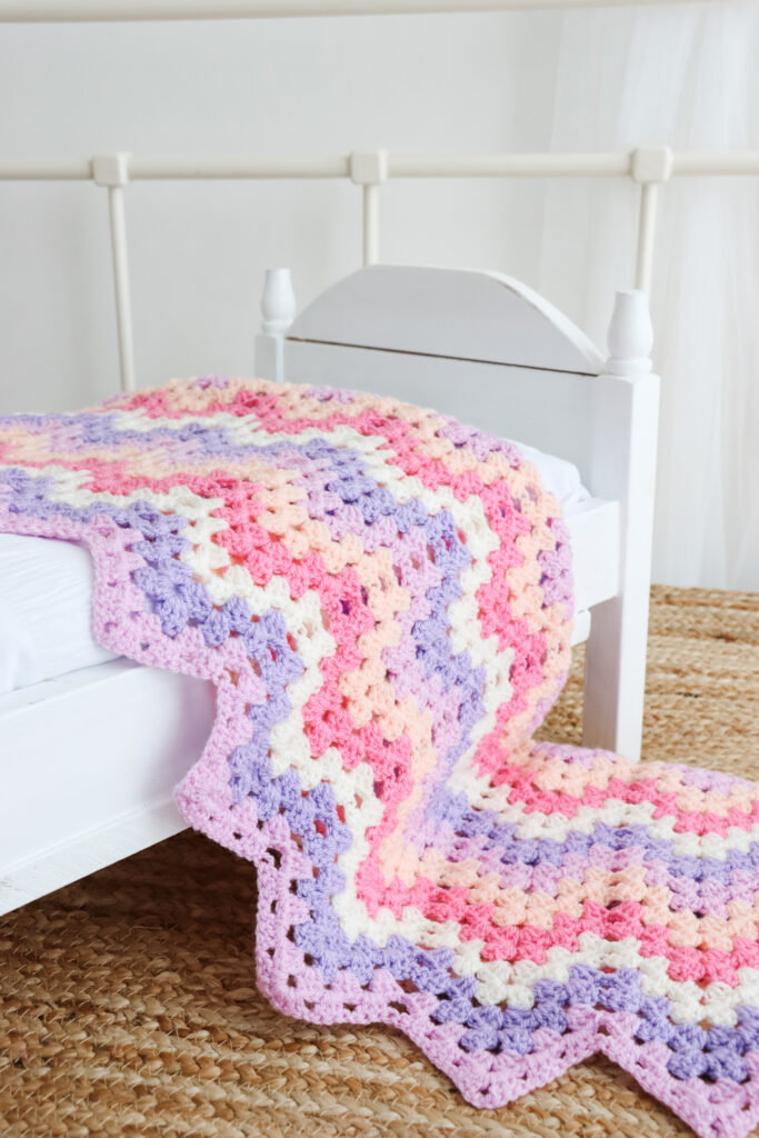 A granny ripple blanket is draped across a bed. The blanket is in shades of lilac, purple, cream, pink and peach.