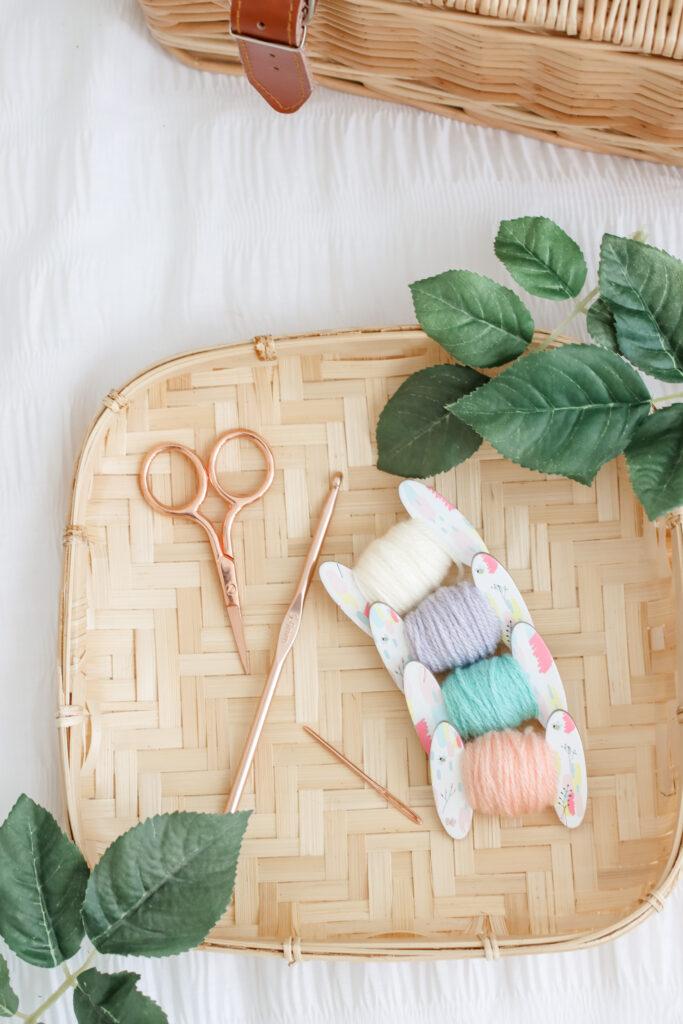 A series of floral bobbins with pastel coloured yarn wrapped around sits on a bamboo plate beside a wicker basket.