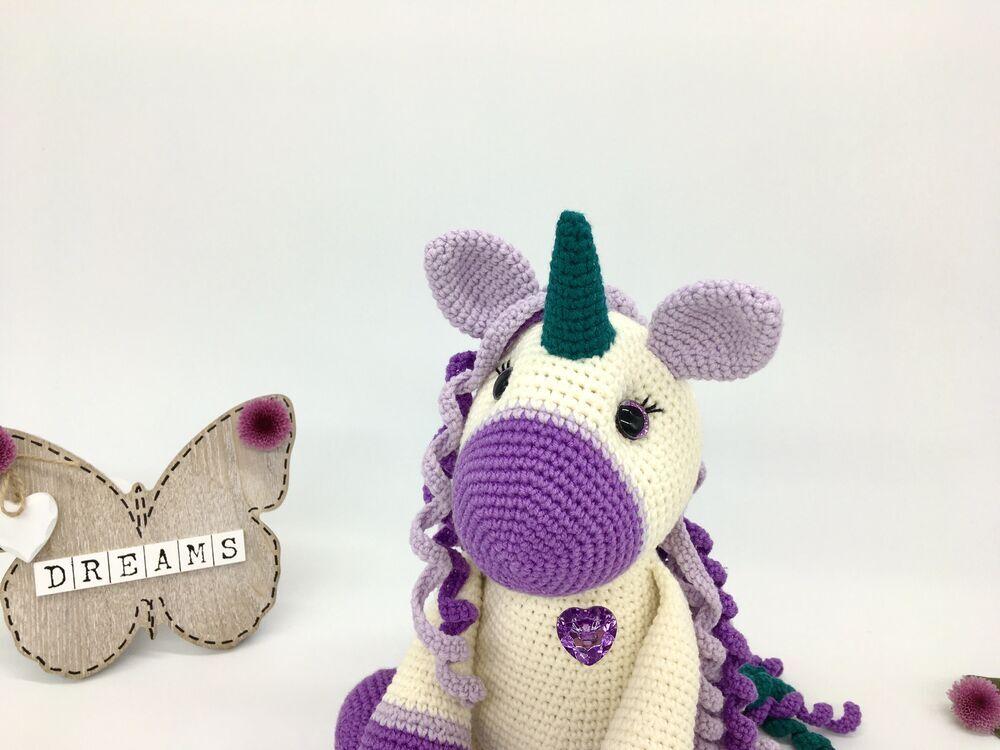 A cream amigurumi unicorn with violet hooves sits on a white background surrounded by purple flowers and a wooden butterfly with the word Dreams on it. The unicorn has curly hair in shades of violet, lilac and team and a teal coloured horn. The crochet toys eyes are large and sparkly and the unicorn has a lilac heart-shaped gem on its chest.