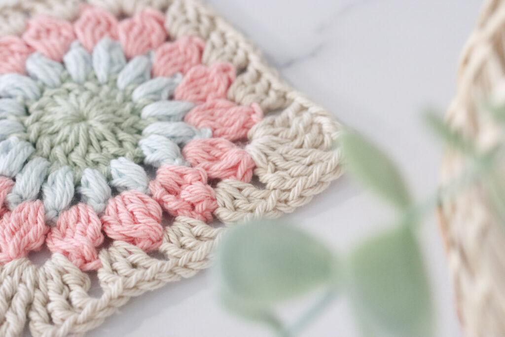 a sunburst granny square lay on a marble backdrop at a slight angle with foliage in the bottom right side corner of the image. The sunburst granny is made from pale green, blue, pink and beige. 
