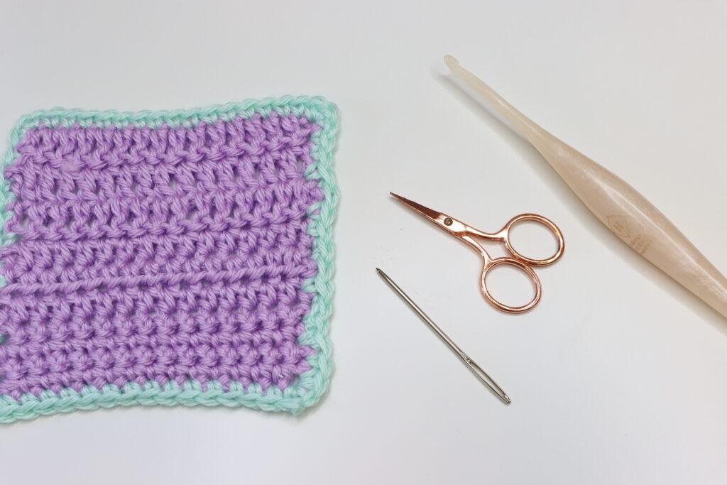 A corner of crochet showing the Camel Stitch border lies on a white background. The swatch is made from lilac and aqua yarn. Beside it are a darning needle, rose gold scissors and a pearlescent crochet hook. 