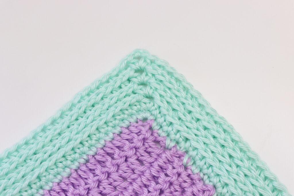 A corner of crochet showing the Camel Stitch border lies on a white background. The swatch is made from lilac and aqua yarn.