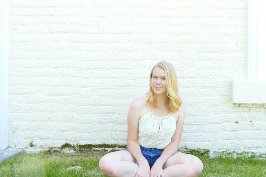 A blonde woman sits cross legged on grass against a white brick wall. She is wearing jean shorts and a white crochet tank top.