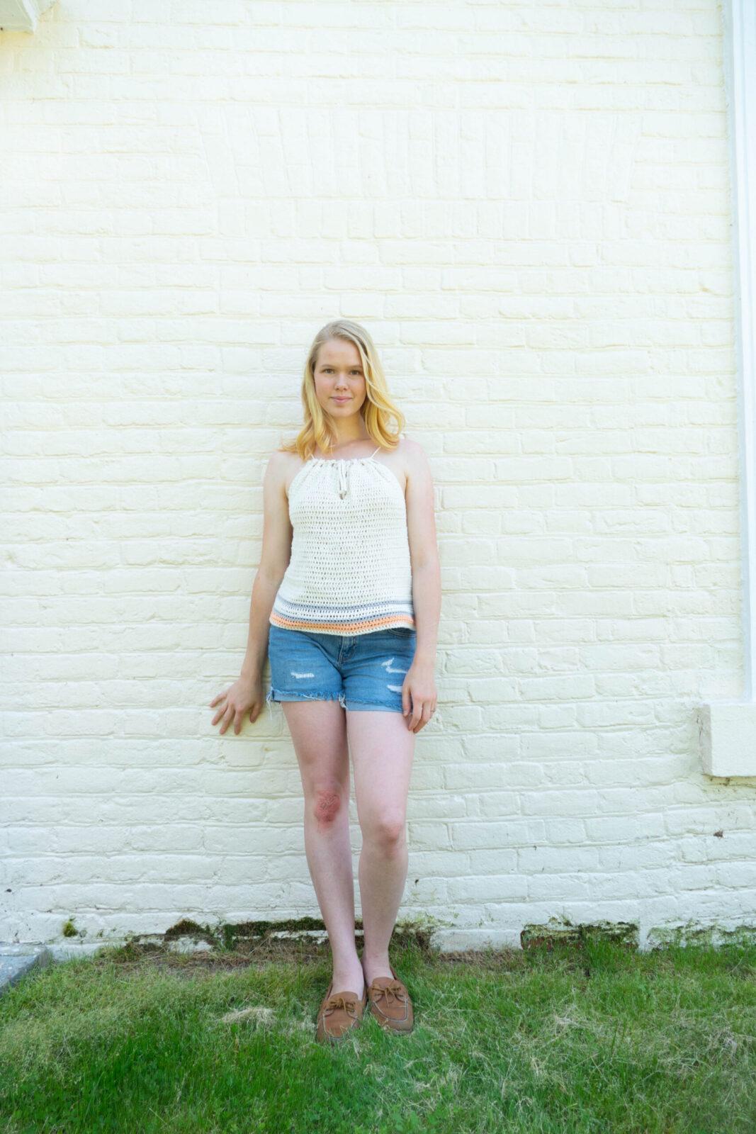 A blonde woman stands against a white brick wall. She is wearing jean shorts and a white crochet tank top.