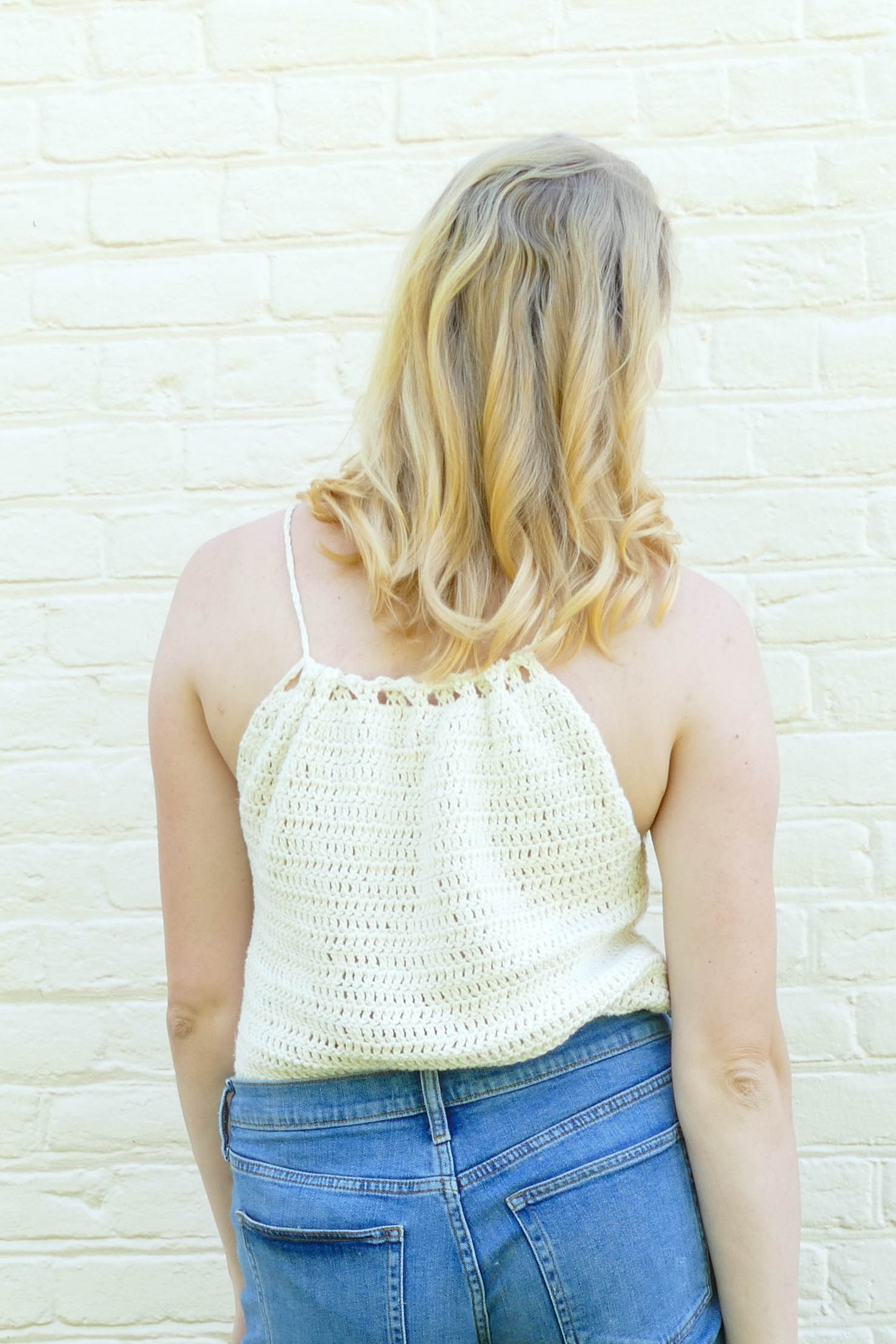 A blonde woman stands against a white brick wall with her back to the camera. She is wearing jean shorts and a white crochet tank top.